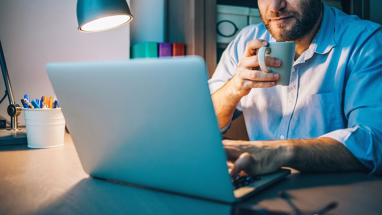 man drinking coffee using laptop at desk with lamp