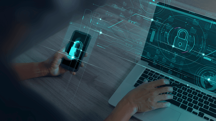 hand holding iphone and another hand typing on open laptop with cybersecurity lock symbols